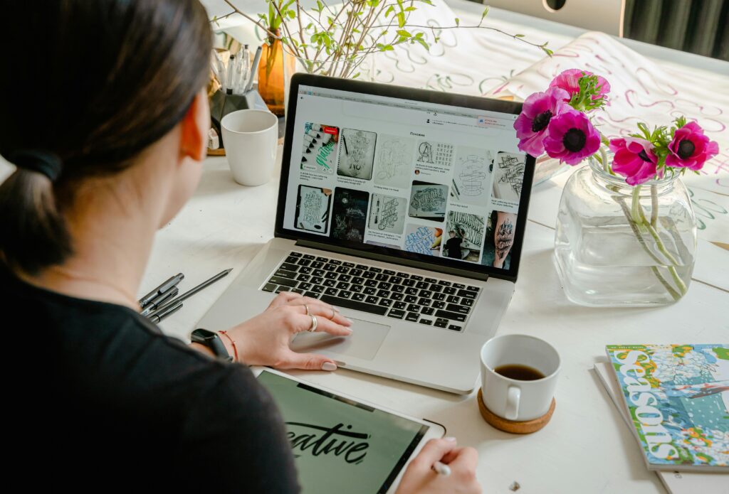 Frau am Laptop ist von hinten zu sehen, während sie arbeitet. Pinkfarbene Blumen stehen in einer Vase als Tischdekoration.