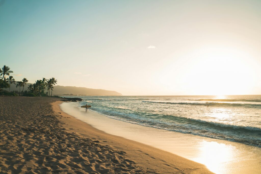 Strand und Meer, In der Ferne steht ein Surfer mit seinem Surfbrett am Strand. Die Sonne spiegelt sich im Meer.